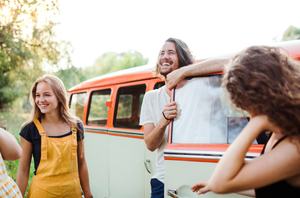 A group of young friends standing by a retro minivan on a roadtrip through countryside.