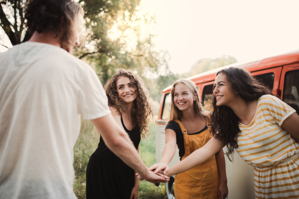 A group of young friends standing by a retro minivan on a roadtrip through countryside, putting hands together.