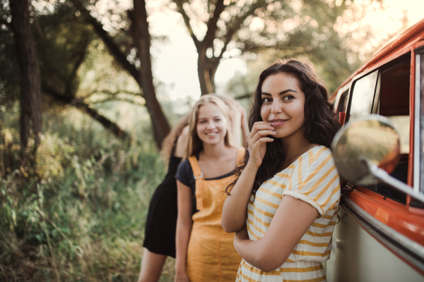 A group of young girl friends standing by a retro minivan on a roadtrip through countryside.