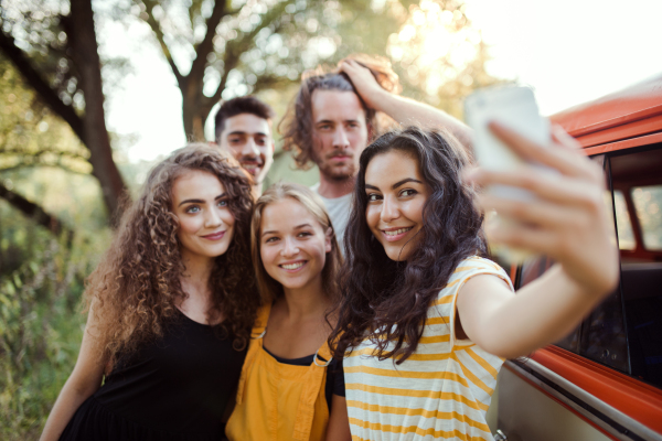 A group of young friends with smartphone on a roadtrip through countryside, taking selfie.