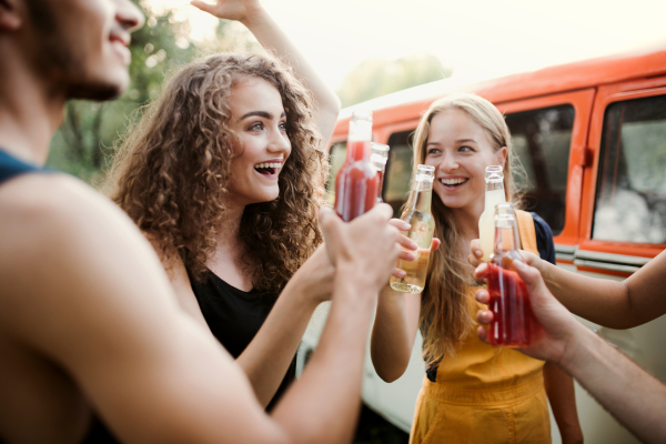 A group of young friends with drinks standing outdoors on a roadtrip through countryside, clinking bottles.