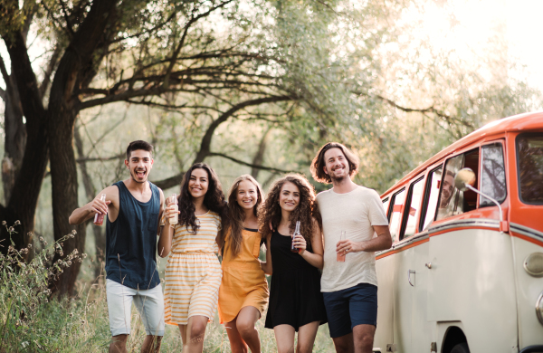 A group of young friends with drinks standing outdoors on a roadtrip through countryside, holding bottles.