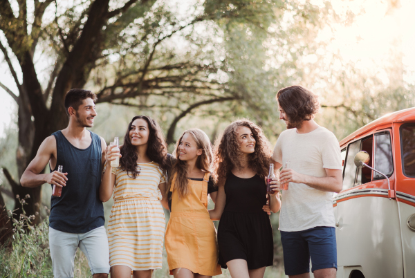 A group of young friends walking by a retro minivan on a roadtrip through countryside, holding bottles.