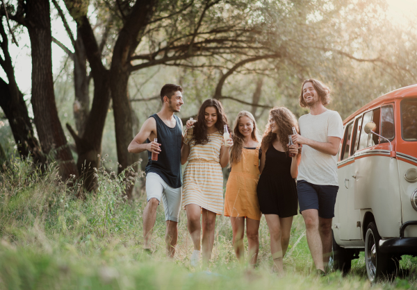 A group of young friends walking by a retro minivan on a roadtrip through countryside, holding bottles.