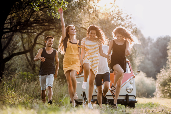 A group of young friends walking in front of a retro minivan on a roadtrip through countryside, jumping.