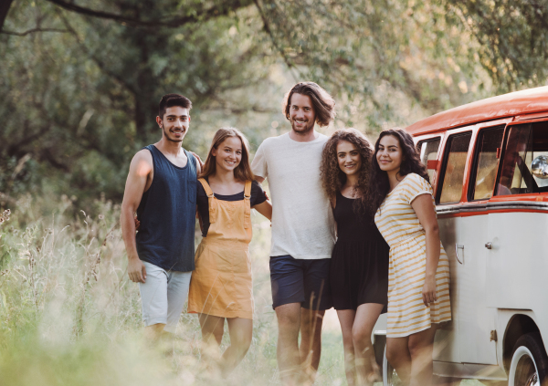 A group of young friends standing by a retro minivan on a roadtrip through countryside.