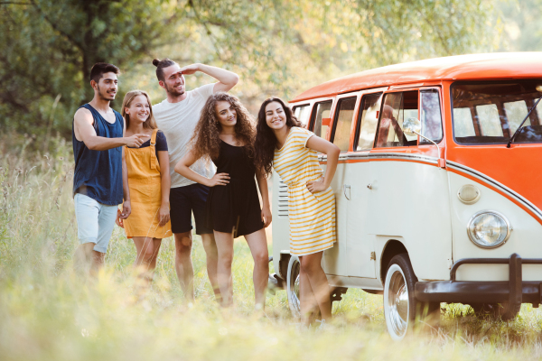 A group of young friends standing by a retro minivan on a roadtrip through countryside.