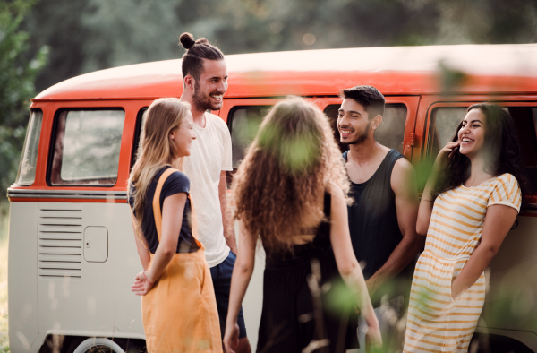 A group of young friends standing by a retro minivan on a roadtrip through countryside, laughing.