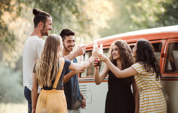 A group of young friends with drinks standing outdoors on a roadtrip through countryside, clinking bottles.