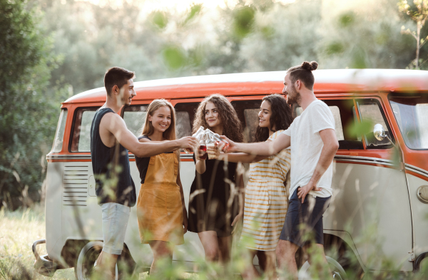 A group of young friends with drinks standing outdoors on a roadtrip through countryside, clinking bottles.