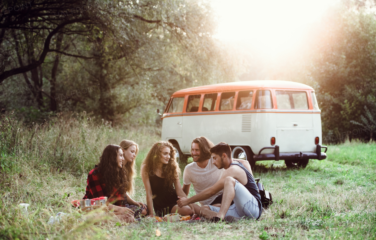 A group of friends sitting on ground by a retro minivan on a roadtrip through countryside, having picnic.