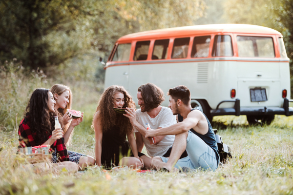 A group of friends sitting on ground by a retro minivan on a roadtrip through countryside, having picnic.