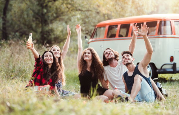 A group of young friends with drinks sitting on grass in front of a retro minivan on a roadtrip through countryside.