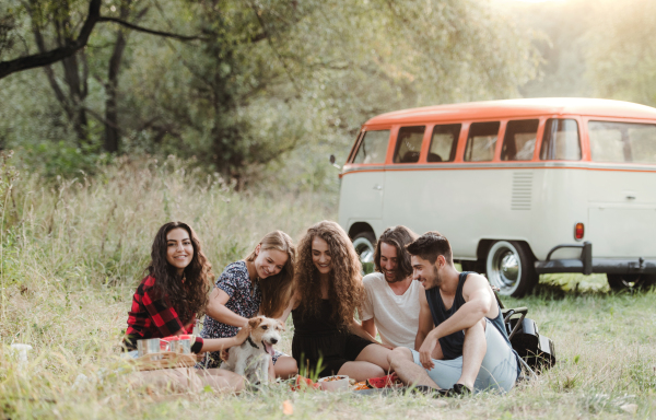 A group of young friends with a dog sitting on grass in front of a retro minivan on a roadtrip through countryside.