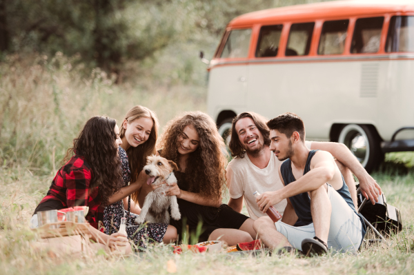 A group of young friends with a dog sitting on grass in front of a retro minivan on a roadtrip through countryside.