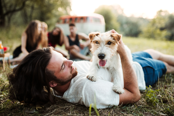 A group of young friends with a dog sitting on grass in front of a retro minivan on a roadtrip through countryside.