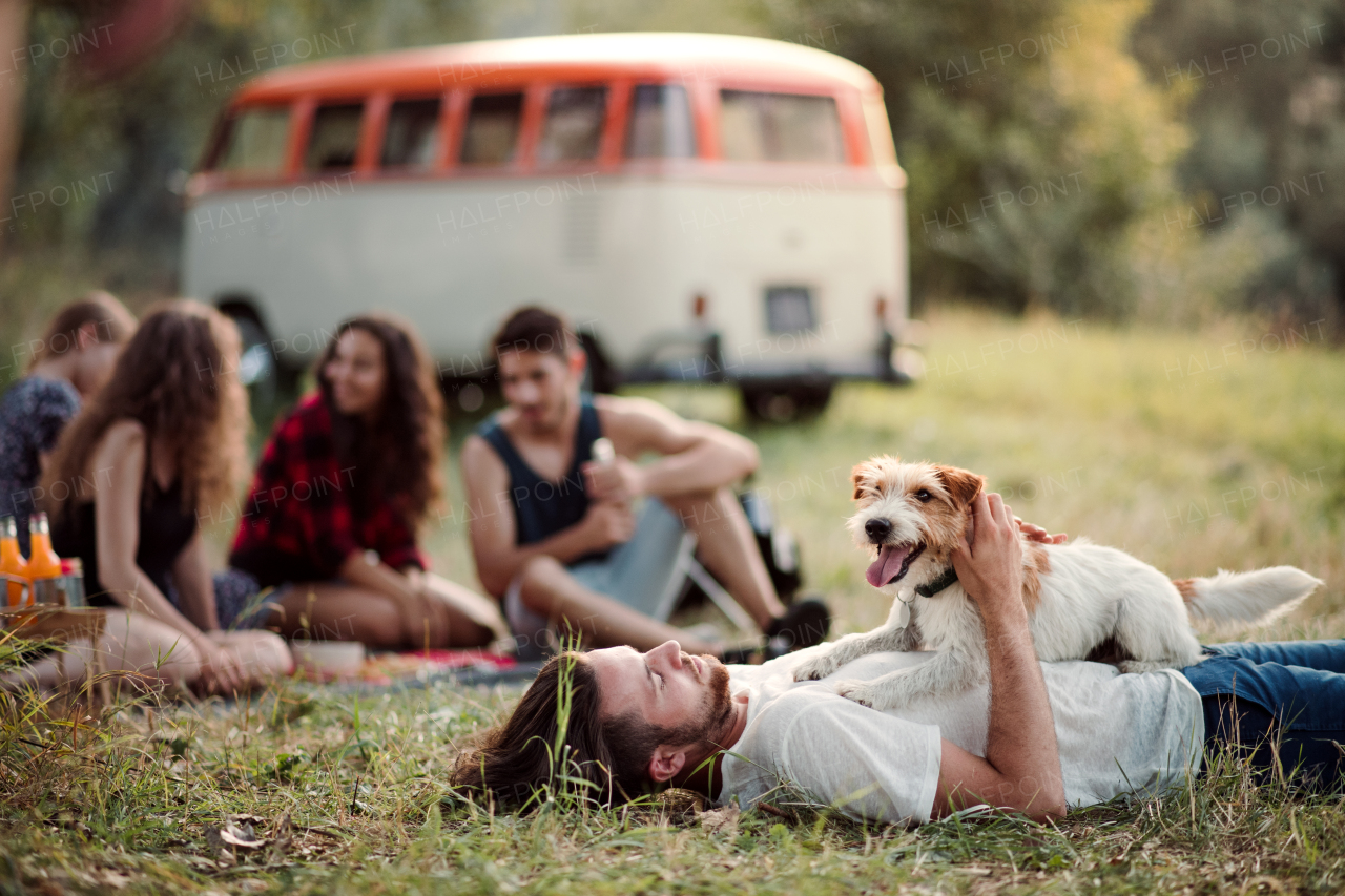 A group of young friends with a dog sitting on grass in front of a retro minivan on a roadtrip through countryside.