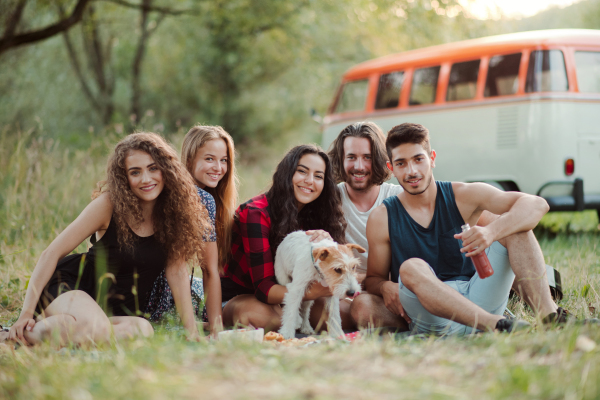 A group of young friends with a dog sitting on grass in front of a retro minivan on a roadtrip through countryside.