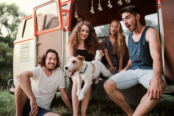 A group of young friends with a dog by a retro minivan on a roadtrip through countryside.