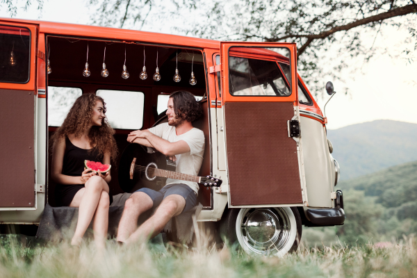 A young couple with guitar and watermelon outdoors on a roadtrip through countryside, talking.