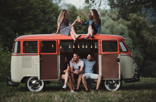 A group of friends at dusk outdoors on a roadtrip through countryside, two girls sitting on the roof of a minivan.