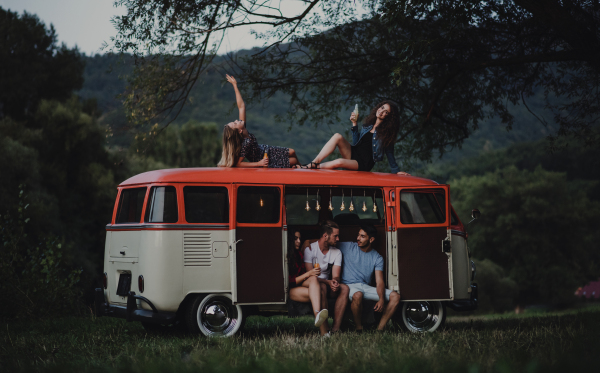 A group of friends at dusk outdoors on a roadtrip through countryside, two girls sitting on the roof of a minivan.