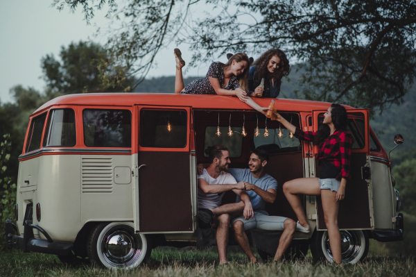 A group of friends at dusk outdoors on a roadtrip through countryside, two girls sitting on the roof of a minivan.