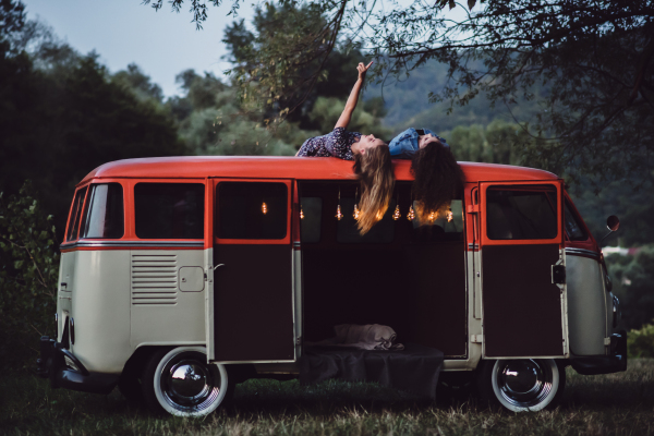 Two girl friends at dusk outdoors on a roadtrip through countryside, having fun on the roof of a minivan.