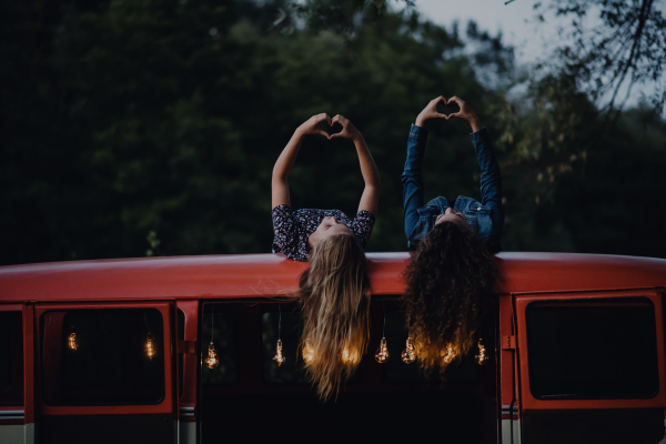 Two girl friends at dusk outdoors on a roadtrip through countryside, having fun on the roof of a minivan.