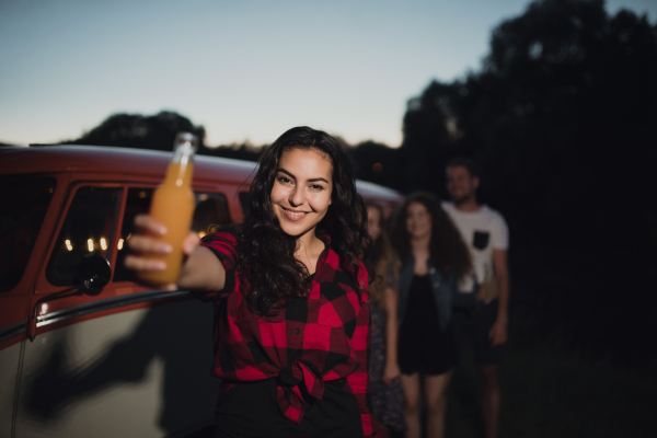 A group of young friends with drinks standing outdoors at dusk on a roadtrip through countryside, holding bottles.