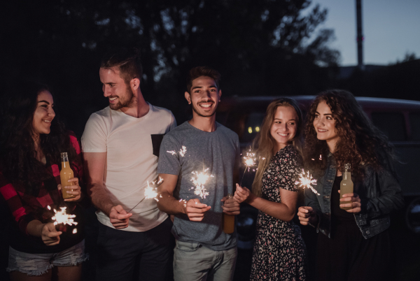 A group of young friends with sparklers standing outdoors on a roadtrip through countryside at dusk.