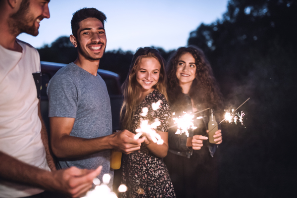 A group of young friends with sparklers standing outdoors on a roadtrip through countryside at dusk.