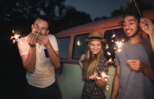 A group of young friends with sparklers standing outdoors on a roadtrip through countryside at dusk.