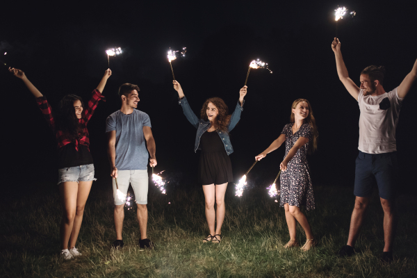 A group of young friends with sparklers standing outdoors on a roadtrip through countryside at dusk.