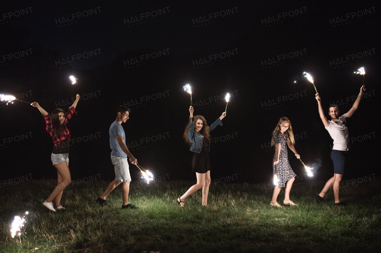 A group of young friends with sparklers standing outdoors on a roadtrip through countryside at dusk.