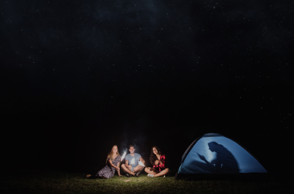 Friends with sparklers outdoors at dusk and one with smartphone in tent, concept of technology addiction versus adventure experience. Copy space.