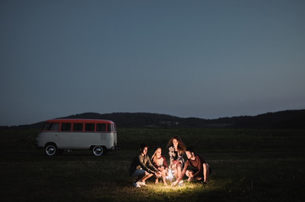 A group of young friends with sparklers standing outdoors on a roadtrip through countryside at dusk. Copy space.