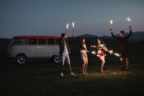 A group of young friends with sparklers standing outdoors on a roadtrip through countryside at dusk.