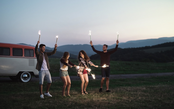 A group of young friends with sparklers standing outdoors on a roadtrip through countryside at dusk.