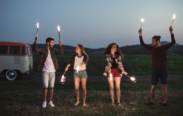 A group of young friends with sparklers standing outdoors on a roadtrip through countryside at dusk.