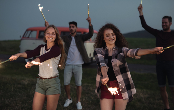 A group of young friends with sparklers standing outdoors on a roadtrip through countryside at dusk.