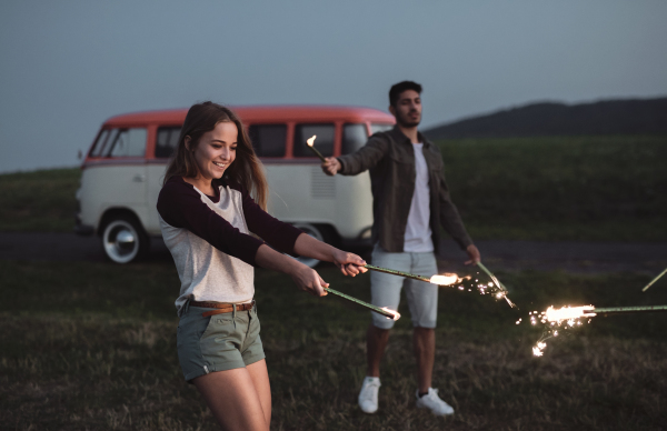 A group of young friends with sparklers standing outdoors on a roadtrip through countryside at dusk.