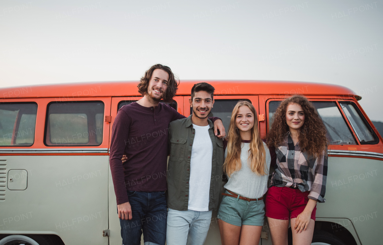 A portrait of group of young friends standing by a retro minivan on a roadtrip through countryside.