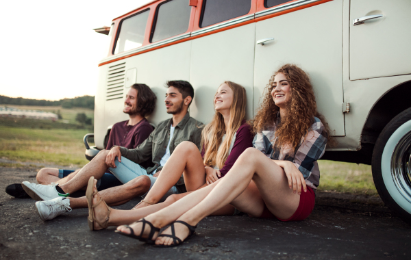 A group of young friends on a roadtrip through countryside, sitting by a retro minivan.