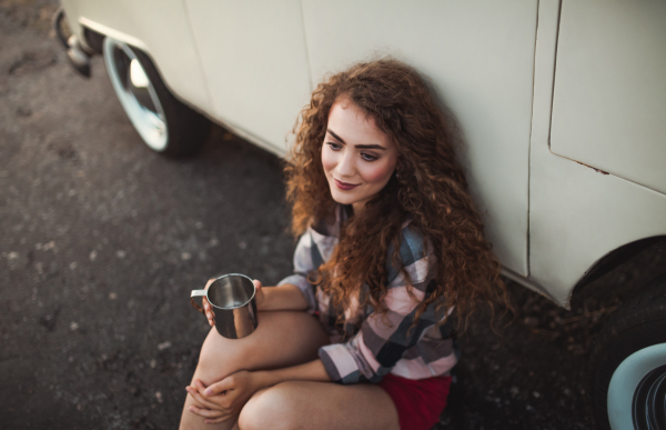 A cheerful young girl sitting by a car on a roadtrip through countryside.