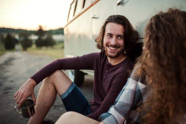 A young couple on a roadtrip through countryside, sitting by retro minivan.