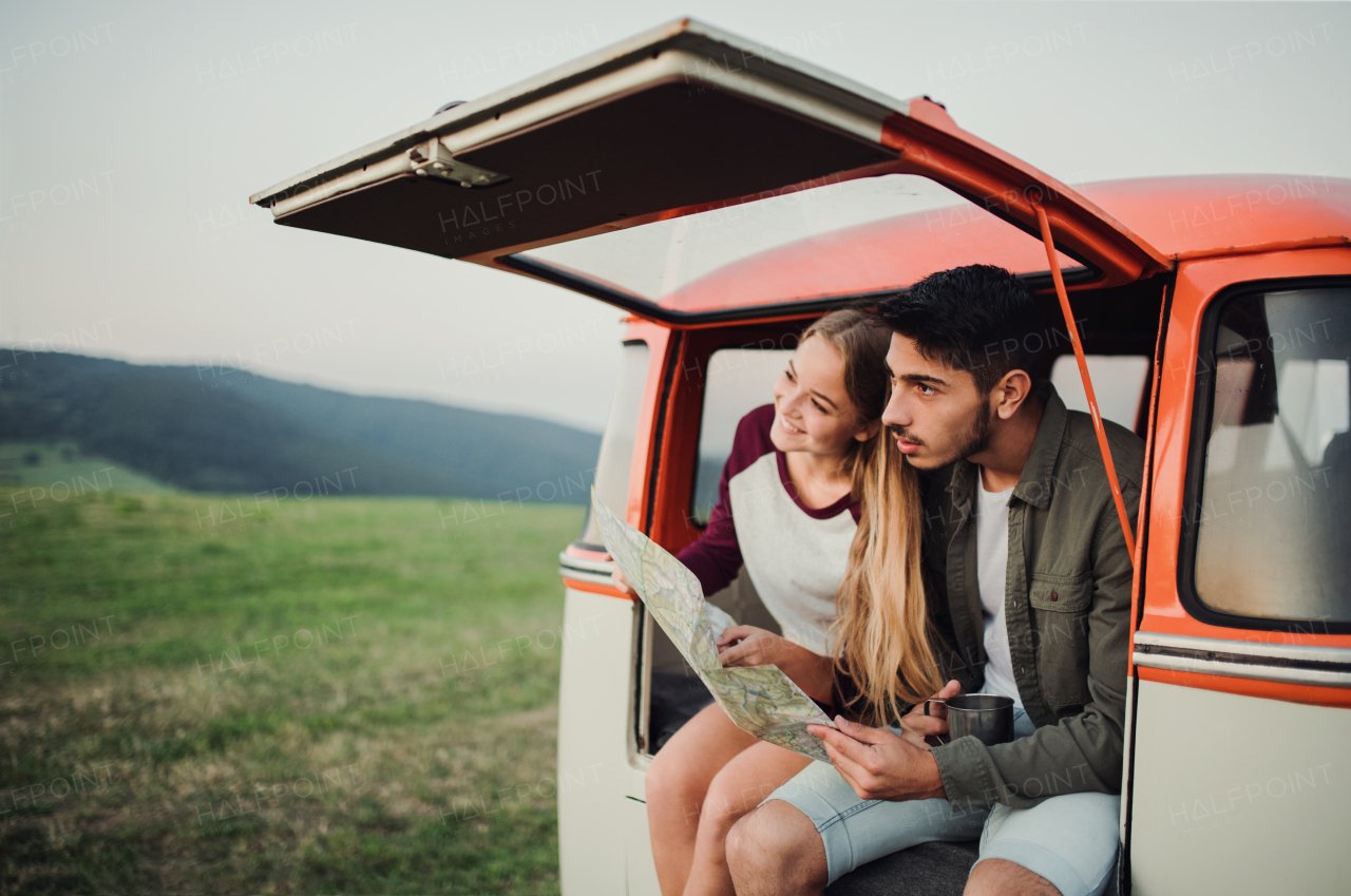 A young couple on a roadtrip through countryside, sitting in retro minivan and looking at a map.