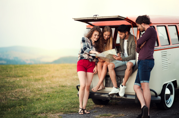 A group of cheerful young friends with a retro minivan on a roadtrip through countryside, looking at map.