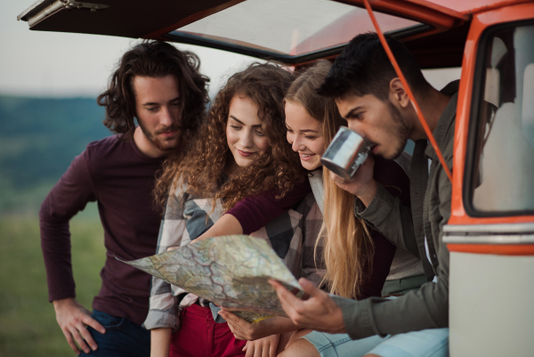 A group of cheerful young friends with a retro minivan on a roadtrip through countryside, looking at map.