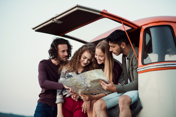 A group of cheerful young friends with a retro minivan on a roadtrip through countryside, looking at map.